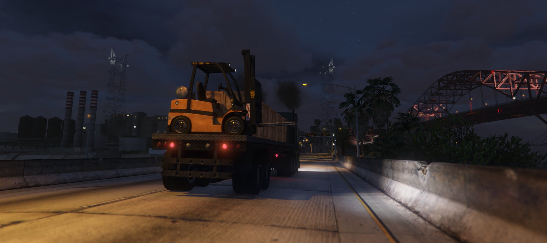 A flatbed truck carrying a forklift along a lit highway at night, refinery and bridge in the distance